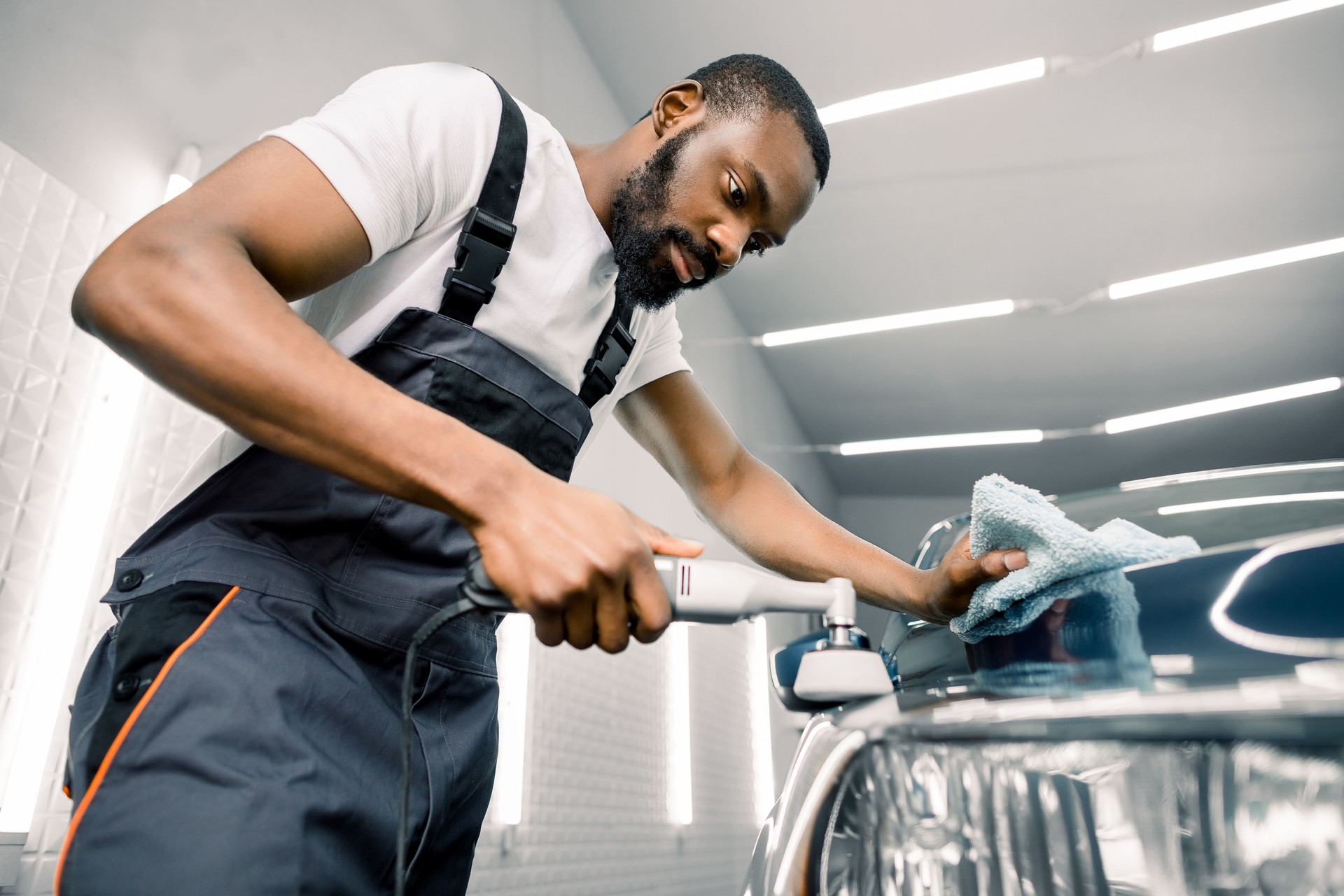 Car detailing polishing process. Handsome concentrated African man worker wearing gray overalls, polishing car lights with orbital polisher in auto detailing service.