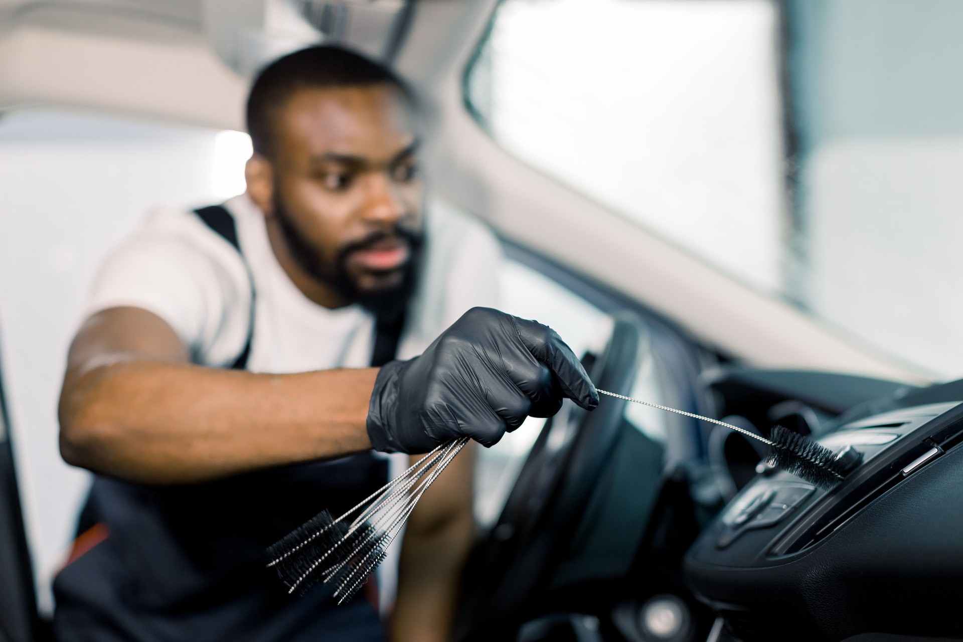 Car detailing and cleaning concept. Young African male worker, wearing uniform and black rubber protective gloves, cleaning car interior with set of black brushes