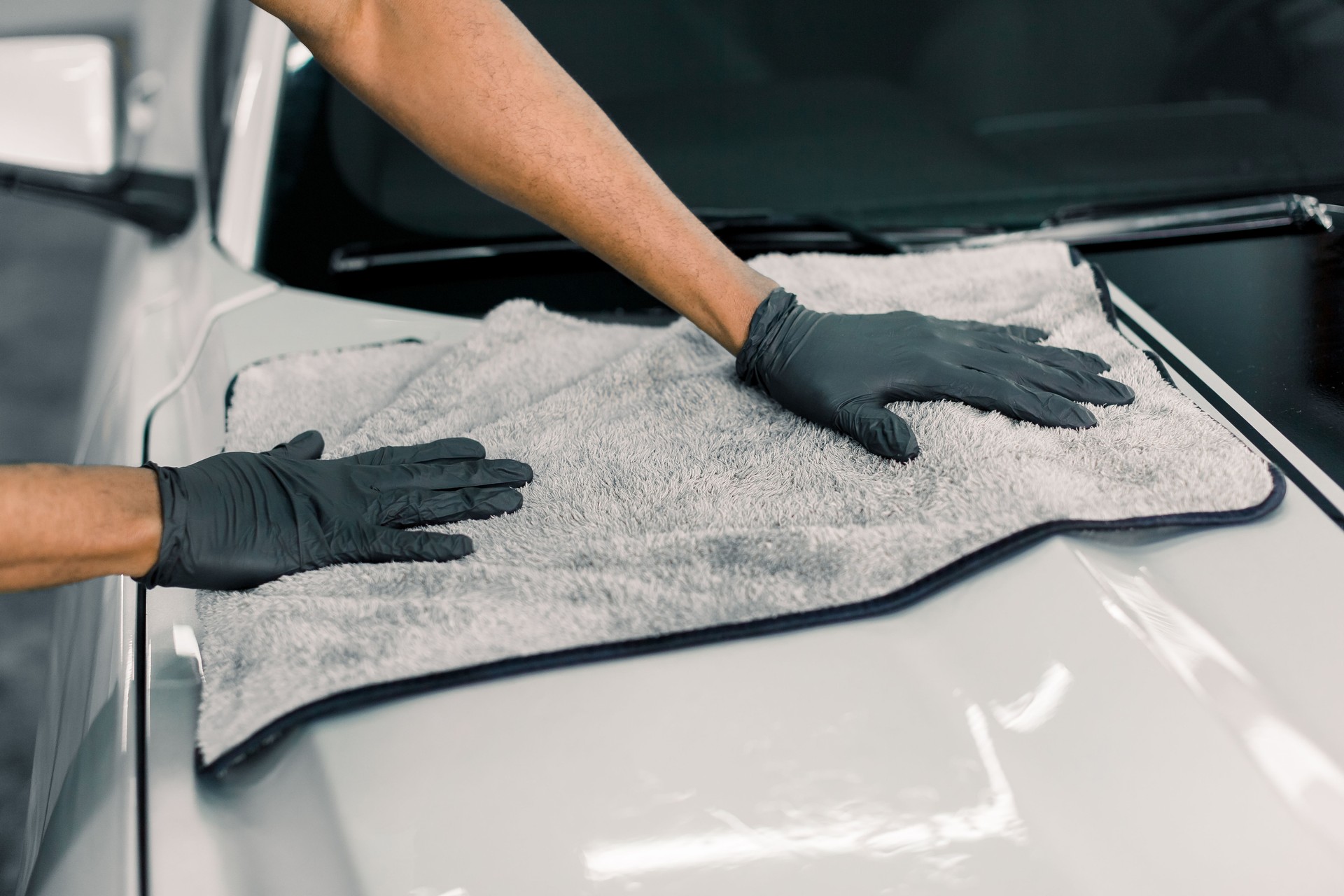 Car detailing, wash and cleaning concept. Cropped image of hands of male professional car wash worker in black rubber gloves, holding the gray microfiber and polishing the car hood of luxury white car
